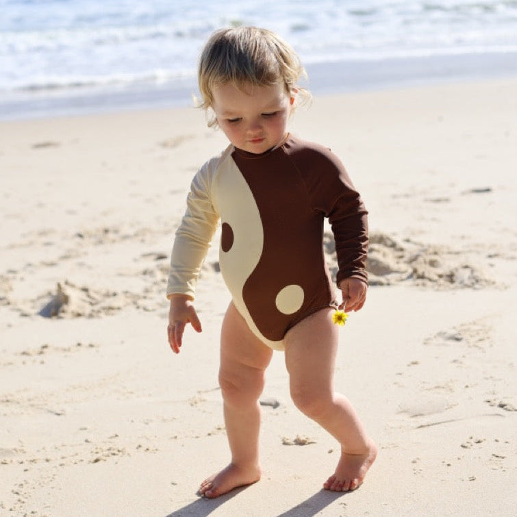 Child in a brown and white yin yang swimsuit standing on a sandy beach with ocean in the background